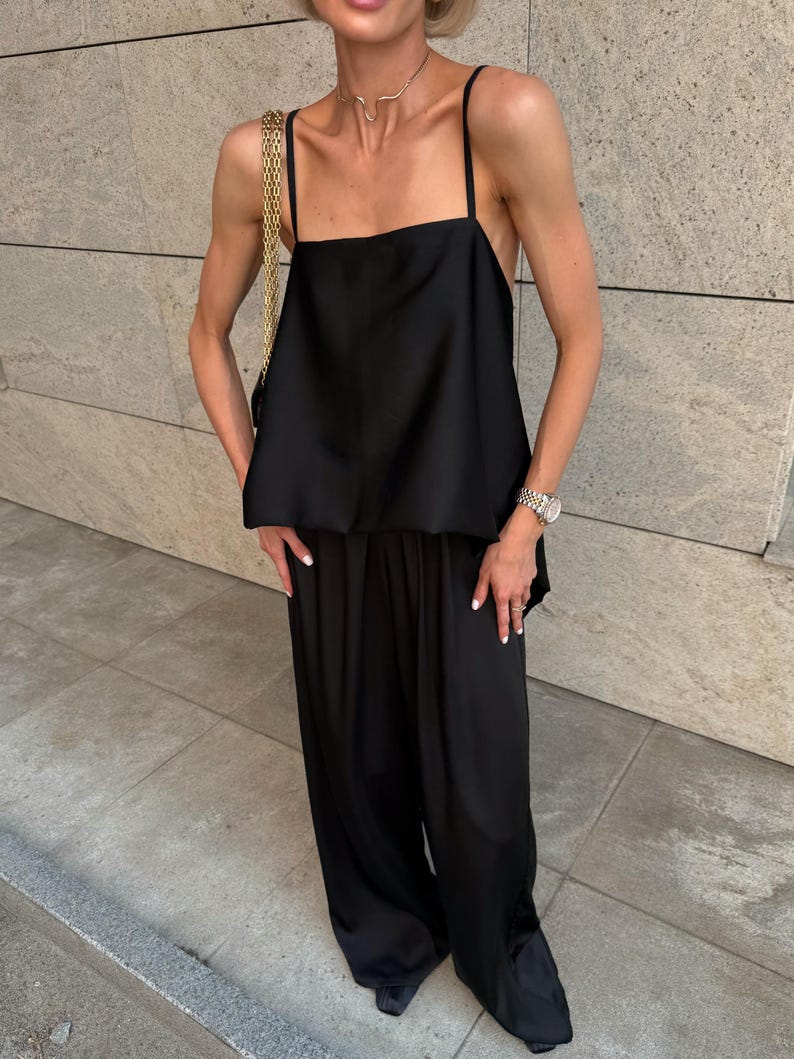 以下が含まれることがあります： 黒いサテンのキャミソールトップ、スパゲッティストラップ、お揃いのワイドレッグパンツ。ゴールドチェーンストラップバッグが肩にかかっています。この服装は、ゴールドのネックレスとシルバーの時計で装飾されています。この服装はモノクロのアンサンブルです。