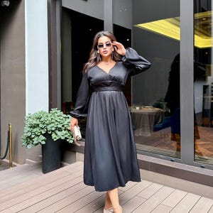 May include: A black midi dress with long sleeves and a V-neck. The dress features a cinched waist and a flared skirt. The person is wearing sunglasses and carrying a small white clutch. The shoes are pale with a bow. The background includes a building and plants.