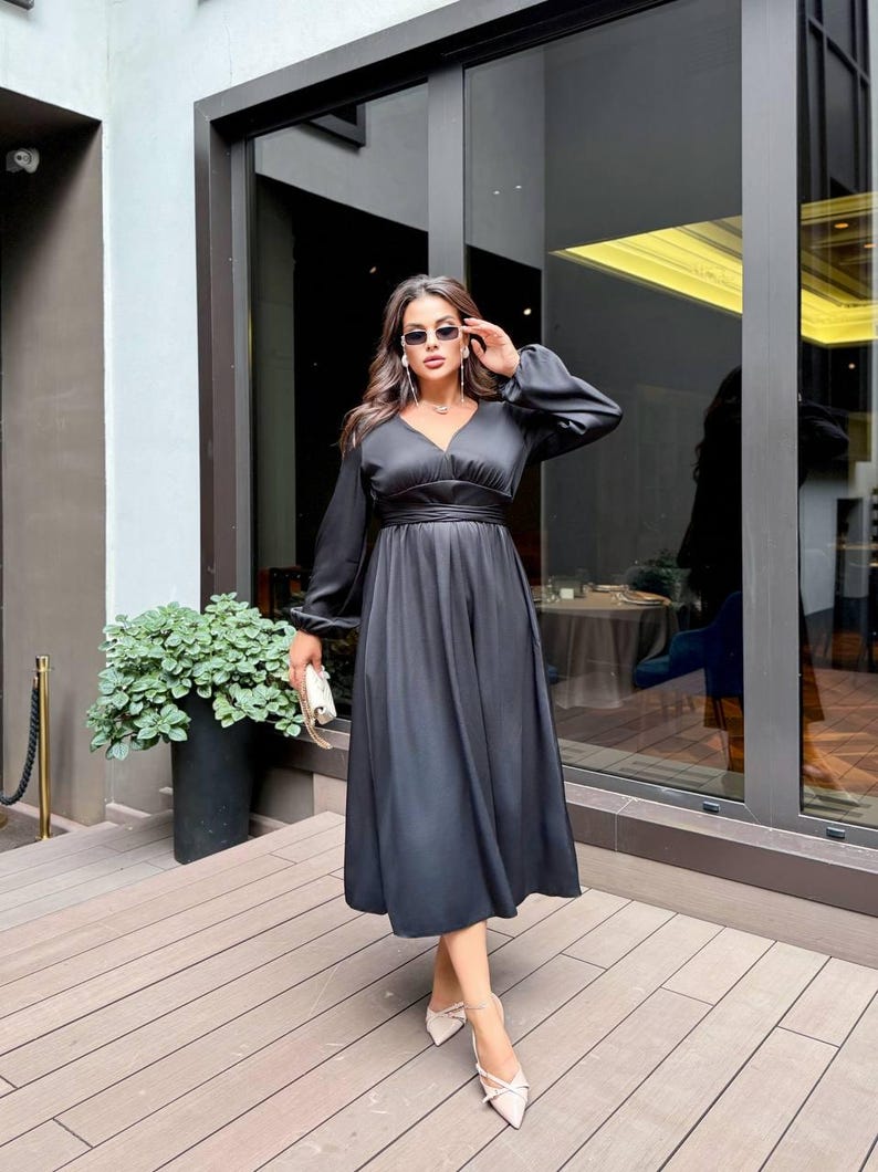 May include: A black midi dress with long sleeves and a V-neck. The dress features a cinched waist and a flared skirt. The person is wearing sunglasses and carrying a small white clutch. The shoes are pale with a bow. The background includes a building and plants.