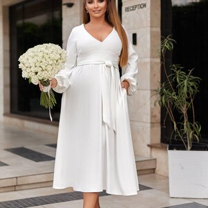 May include: A white midi dress with long sleeves and a V-neck. The dress has a tie waist and pockets. A large bouquet of white roses is held. The person is wearing light pink heels. The word "RECEPTION" is visible in the background.
