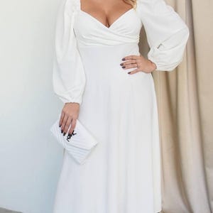 May include: A white midi dress with long, puffy sleeves and a sweetheart neckline. The dress is paired with white heels and a white clutch. The clutch has a black logo. The dress is a solid color and appears to be made of a soft fabric.