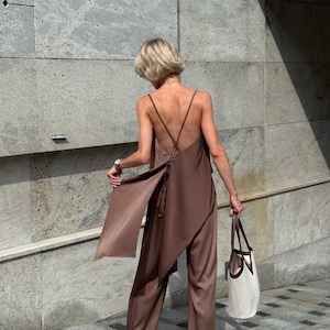 以下が含まれることがあります： ブラウンのサテンジャンプスーツ。流れるような非対称のトップスとワイドパンツ。トップスは背中で交差する細いストラップが特徴です。茶色のハンドルが付いた白いトートバッグが右手で持たれています。