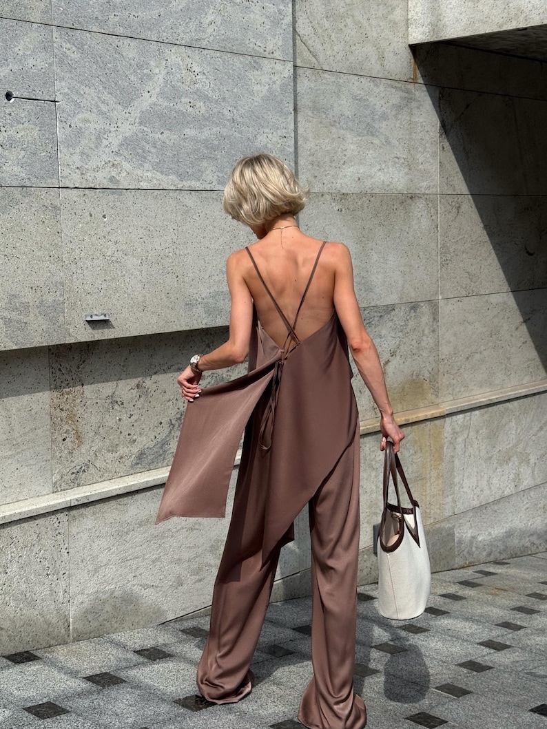 以下が含まれることがあります： ブラウンのサテンジャンプスーツ。流れるような非対称のトップスとワイドパンツ。トップスは背中で交差する細いストラップが特徴です。茶色のハンドルが付いた白いトートバッグが右手で持たれています。