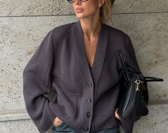 May include: A dark gray cardigan with a V-neck, button closure, and front pockets. The cardigan is paired with dark wash jeans and a black handbag. The person is wearing black-framed glasses.