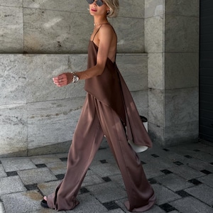 以下が含まれることがあります： ブラウンのサテンキャミソールと、お揃いのワイドパンツのコーディネート。背中が大きく開いたデザインで、流れるようなシルエットが特徴です。モデルはサングラスとサンダルを着用しています。単色で洗練された印象を与えます。