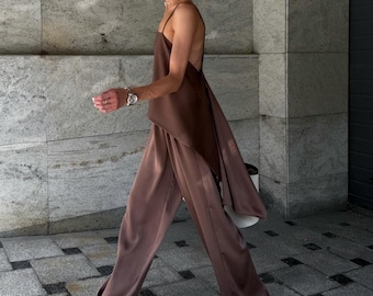 May include: A brown satin top with spaghetti straps and a flowing back detail, paired with matching wide-leg trousers. The outfit is a solid color, creating a sleek, monochromatic look. The model is wearing sunglasses and sandals.