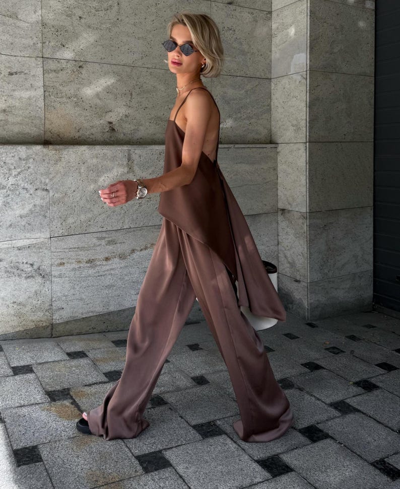 以下が含まれることがあります： ブラウンのサテンキャミソールと、お揃いのワイドパンツのコーディネート。背中が大きく開いたデザインで、流れるようなシルエットが特徴です。モデルはサングラスとサンダルを着用しています。単色で洗練された印象を与えます。