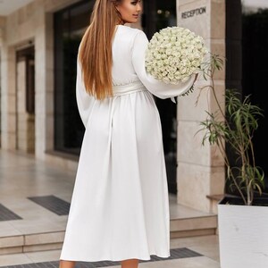 May include: A white, long-sleeved dress with a matching belt. The dress is worn with light pink heels. A large bouquet of white roses is held. The background features a building with the word "RECEPTION".