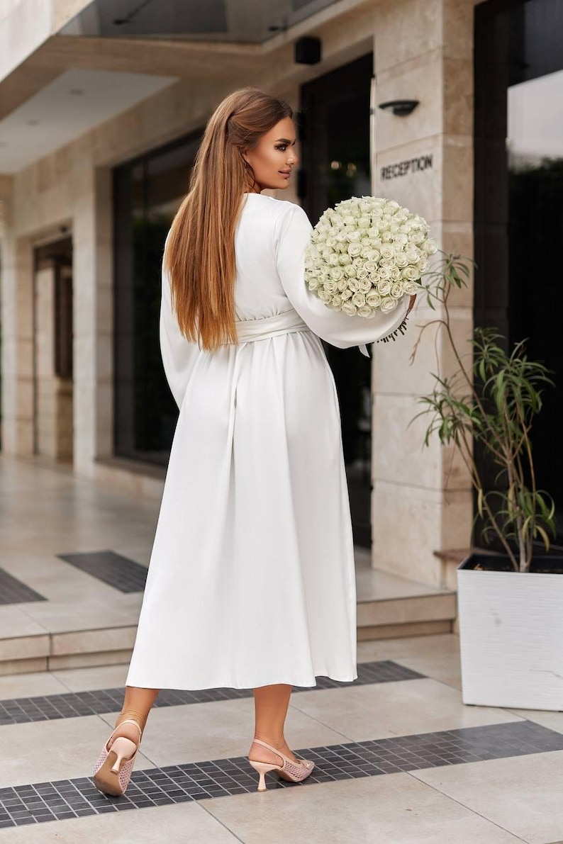 May include: A white, long-sleeved dress with a matching belt. The dress is worn with light pink heels. A large bouquet of white roses is held. The background features a building with the word "RECEPTION".