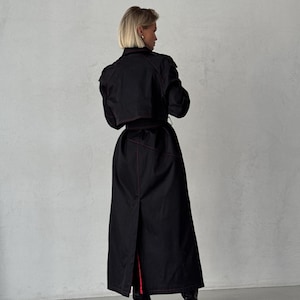 May include: A long black trench coat with red stitching. The coat has a belt and a long slit in the back.