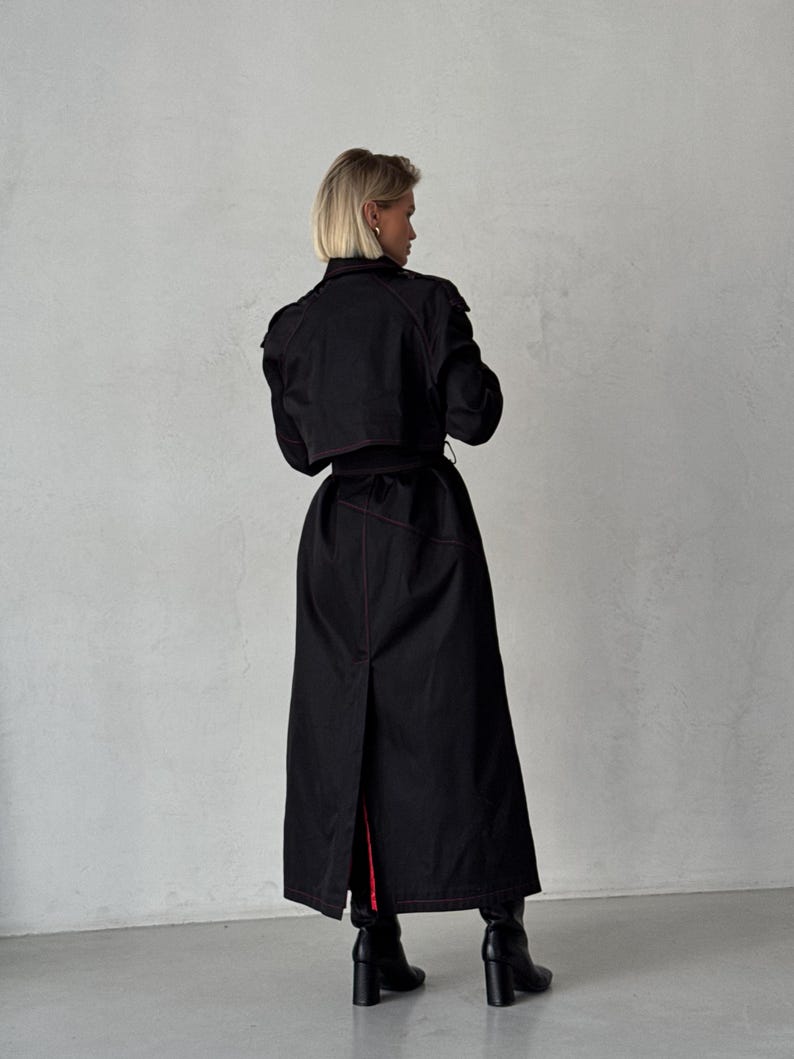 May include: A long black trench coat with red stitching. The coat has a belt and a long slit in the back.