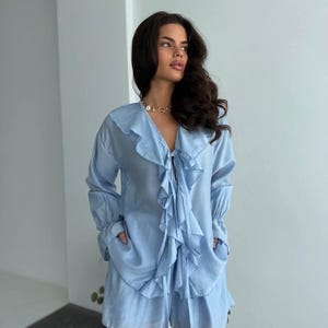 以下が含まれることがあります： フリル付きの長袖トップスとショートパンツが特徴の、ライトブルーのツーピースセット。トップスはVネックで、リボン結びとポケットが付いています。ショートパンツにもポケットが付いています。セットはシルクのような素材で作られています。