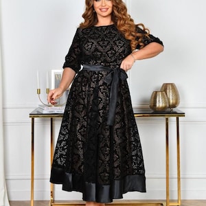 May include: A black lace dress with a satin belt and hem. The dress has short sleeves and a knee-length skirt. The model is wearing red heels. The dress is displayed on a gold table.