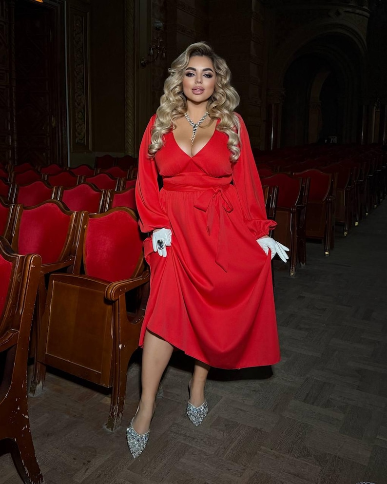 May include: A red midi dress with long sleeves and a wrap-style bodice. The dress is cinched at the waist with a matching belt. The person is wearing white gloves and silver embellished heels. The background features red theatre seats.