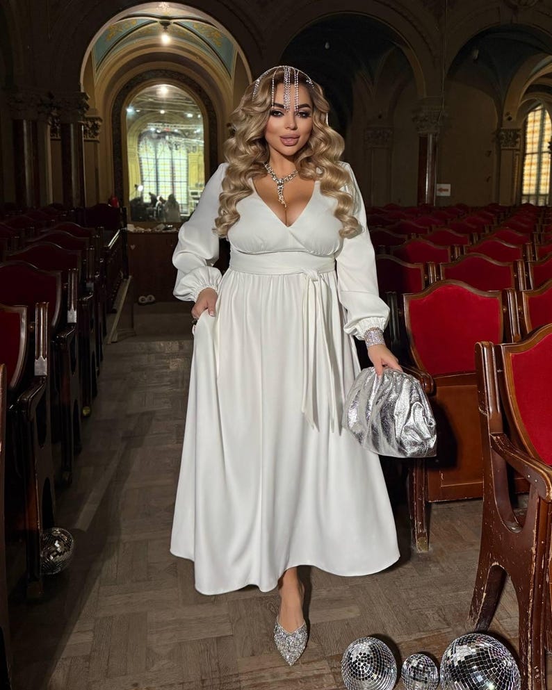 May include: A white long-sleeved dress with a wrap-style bodice and a matching belt. The dress is paired with silver accessories, including a clutch, shoes, and a headpiece. The background features red seating and a wooden floor.