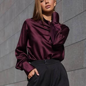 May include: A burgundy satin blouse with long sleeves and a draped neckline. The blouse is paired with black high-waisted trousers. The outfit is simple and elegant, suitable for a variety of occasions.