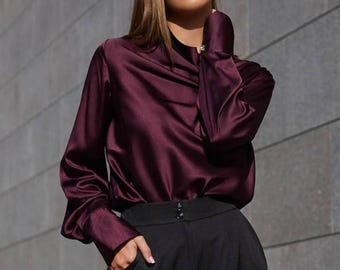 May include: A burgundy satin blouse with long sleeves and a draped neckline. The blouse is paired with black high-waisted trousers. The outfit is simple and elegant, suitable for a variety of occasions.