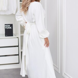 May include: A long, white dress with long sleeves and a tie waist. The dress is paired with white heels. The background includes a white wardrobe and a shelf with books that read "NEW YORK" and "STYLE PARIS".