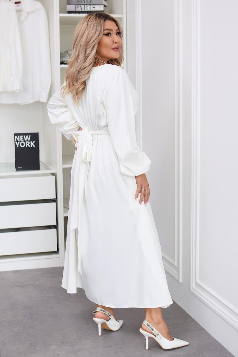 May include: A long, white dress with long sleeves and a tie waist. The dress is paired with white heels. The background includes a white wardrobe and a shelf with books that read "NEW YORK" and "STYLE PARIS".
