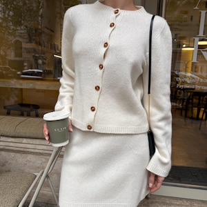 May include: A cream-colored knit two-piece set consisting of a long-sleeved cardigan with a round neckline and seven brown buttons down the front, paired with a matching mini skirt. A black shoulder bag with a thin gold chain strap is worn.