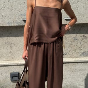 以下が含まれることがあります： 茶色のツーピースの衣装。スクエアネックのキャミソールとワイドパンツで構成されています。シルクのような素材で作られています。クリーム色のボディと茶色のハンドルが付いた大きなトートバッグを持っています。