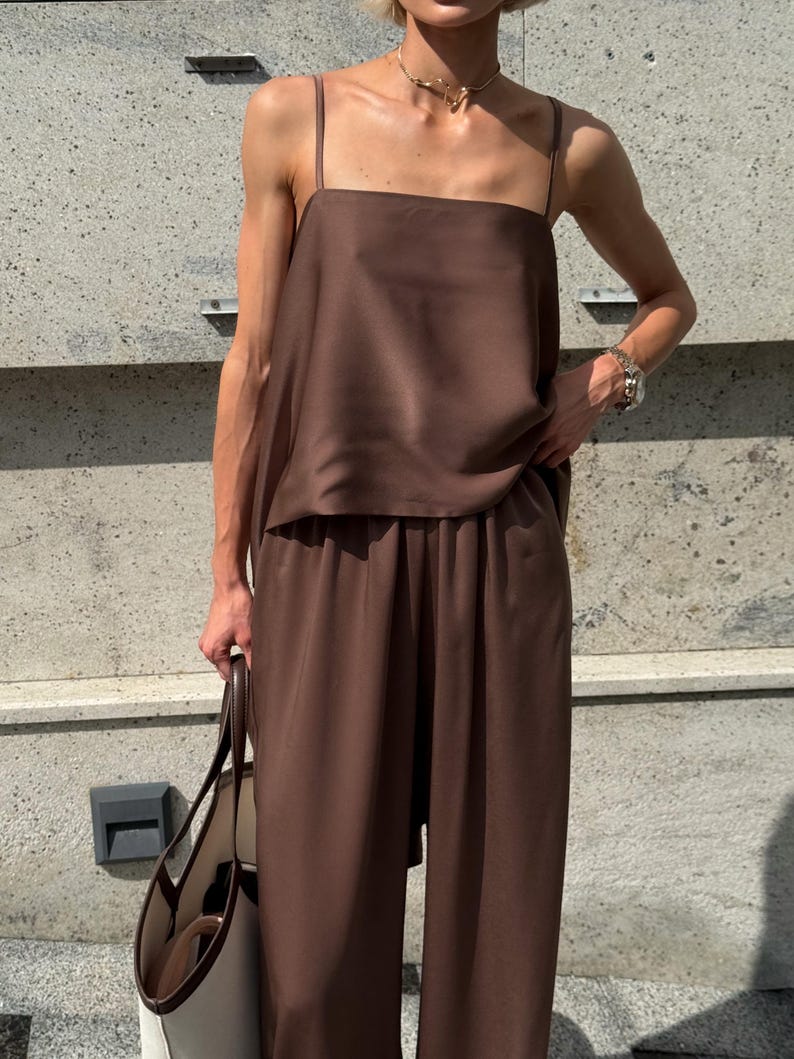 以下が含まれることがあります： 茶色のツーピースの衣装。スクエアネックのキャミソールとワイドパンツで構成されています。シルクのような素材で作られています。クリーム色のボディと茶色のハンドルが付いた大きなトートバッグを持っています。