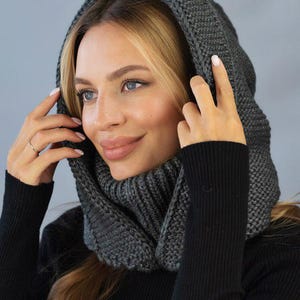 Può includere: Un set di cappuccio e sciarpa in maglia grigio scuro. Il cappuccio è tirato su, incorniciando il viso. La sciarpa avvolge il collo. La persona indossa una maglia nera a maniche lunghe. Lo sfondo è grigio chiaro.