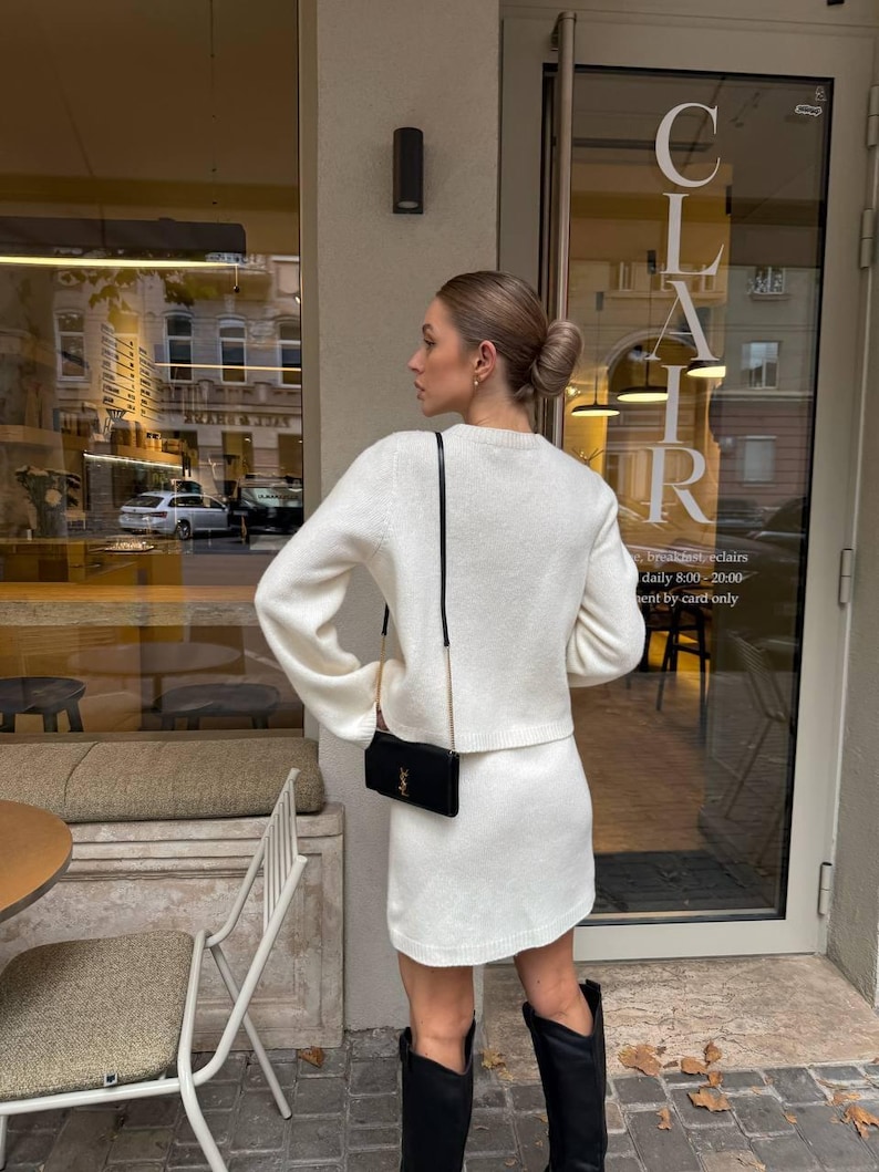 May include: A cream-colored long-sleeve knit sweater and matching knit mini skirt set. The outfit is paired with a black rectangular shoulder bag featuring a gold logo and black knee-high boots. The ensemble is worn with hair in a bun.