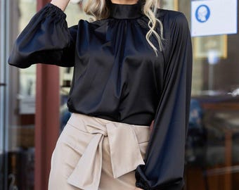 May include: A black satin blouse with long, gathered sleeves and a high neckline. The blouse is paired with a beige skirt featuring a large bow detail at the waist. The outfit is a stylish ensemble.