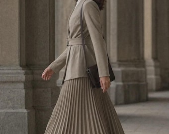 May include: A beige belted blazer with long sleeves and a high collar, worn over a white mock neck top and a matching beige midi-length pleated skirt. A dark brown leather crossbody bag with a thin strap and black ankle boots complete the outfit.