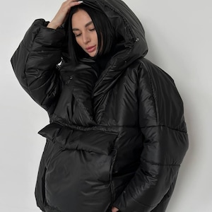 Puede incluir: Una chaqueta acolchada negra con una gran capucha. La chaqueta tiene un ajuste holgado, un gran bolsillo delantero y un cordón en el dobladillo. La modelo lleva la chaqueta. La chaqueta es una prenda de abrigo de invierno.
