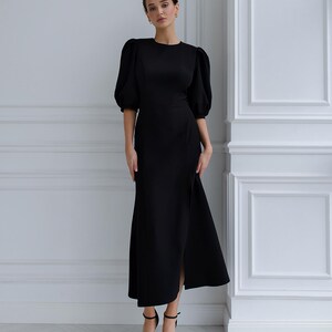 May include: A black dress featuring a round neckline and elbow-length, puffed sleeves. The dress has a side slit and is worn with black high-heeled sandals. The background is a white wall.