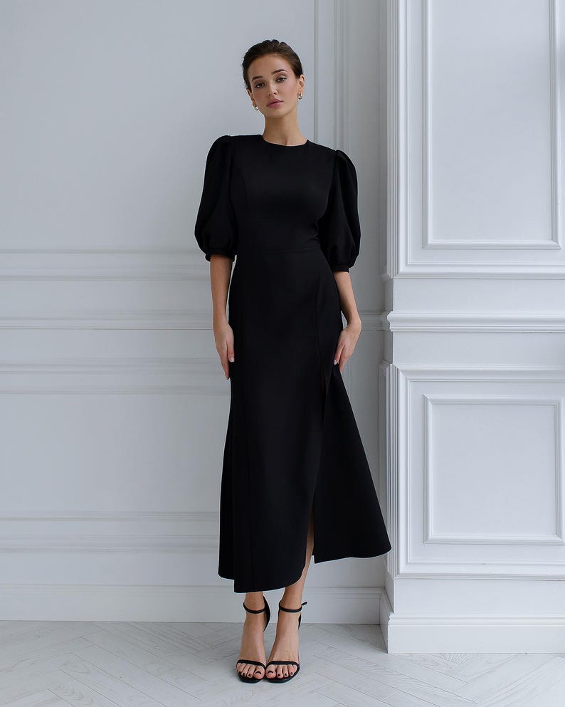 May include: A black dress featuring a round neckline and elbow-length, puffed sleeves. The dress has a side slit and is worn with black high-heeled sandals. The background is a white wall.