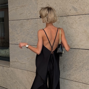 以下が含まれることがあります： 黒いジャンプスーツ、ローバックとスパゲッティストラップ付き。ジャンプスーツは背面に流れるようなレイヤードデザインとワイドレッグパンツが特徴です。ゴールドチェーンストラップ付きの小さな黒いハンドバッグが見えます。