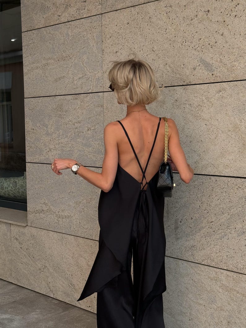 以下が含まれることがあります： 黒いジャンプスーツ、ローバックとスパゲッティストラップ付き。ジャンプスーツは背面に流れるようなレイヤードデザインとワイドレッグパンツが特徴です。ゴールドチェーンストラップ付きの小さな黒いハンドバッグが見えます。