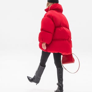Peut inclure: Une doudoune rouge vif oversize à col montant, associée à un legging noir, des bottes hautes en cuir noir à petit talon et un petit sac bandoulière rouge. Un bonnet noir est porté sur la tête, complétant ainsi cette tenue vestimentaire.