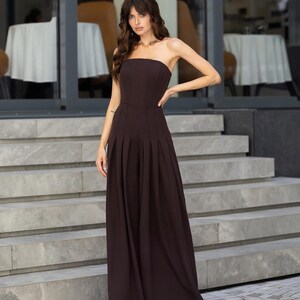 May include: A dark brown strapless jumpsuit featuring a fitted bodice and wide-leg trousers with vertical pleats extending from the waistline to the floor. The garment has a clean, structured silhouette with a straight neckline and no sleeves.