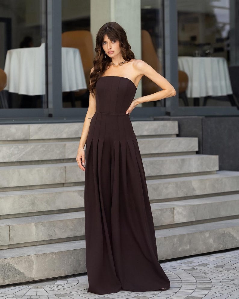 May include: A dark brown strapless jumpsuit featuring a fitted bodice and wide-leg trousers with vertical pleats extending from the waistline to the floor. The garment has a clean, structured silhouette with a straight neckline and no sleeves.