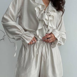 以下が含まれることがあります： 淡いベージュのツーピースパジャマセット。長袖トップスは、フリルカラーと袖口、ネックラインにリボンディテールが特徴です。お揃いのショートパンツは、ドローストリングウエストです。生地は微妙な光沢があります。チャーム付きのゴールドネックレスが見えます。