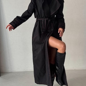 May include: A long black trench coat with red stitching. The coat has a belt and a single button closure. The model is wearing black boots.