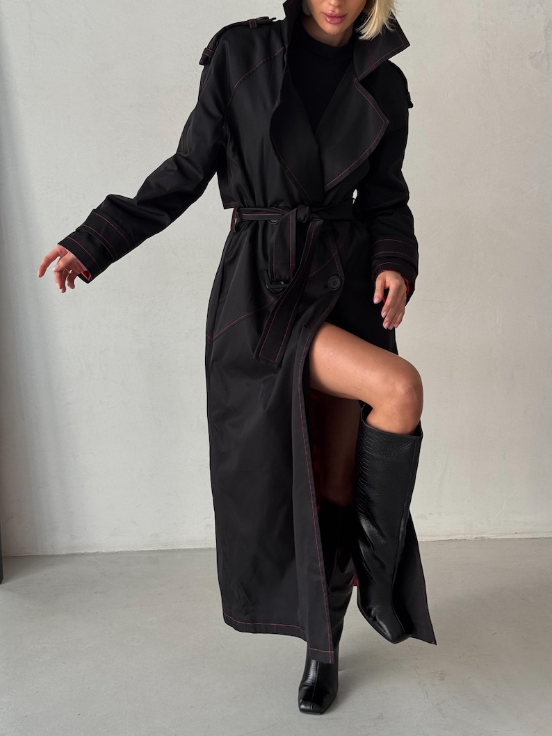 May include: A long black trench coat with red stitching. The coat has a belt and a single button closure. The model is wearing black boots.