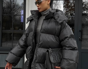 May include: A black puffer jacket with a large hood and a matching black handbag. The jacket has a zip closure and a quilted design. The handbag has a chain strap and a flap closure. The person is wearing sunglasses and a gray turtleneck.