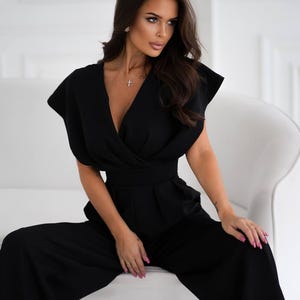 May include: A solid black jumpsuit featuring a deep V-neckline, short wide sleeves, and a fitted waistline that transitions into wide-leg trousers. The garment has a wrap-style bodice with structured shoulders and a clean, minimalist silhouette.