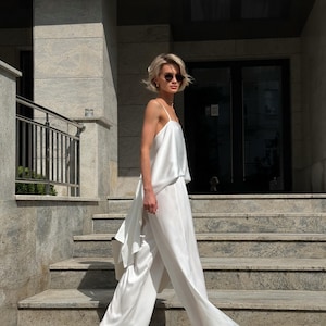 以下が含まれることがあります： 白いサテンのツーピースの衣装。キャミソールとワイドパンツで構成されています。短いブロンドの髪とサングラスをかけた人が、石段を歩いています。この衣装は、流れるようなエレガントな外観をしています。
