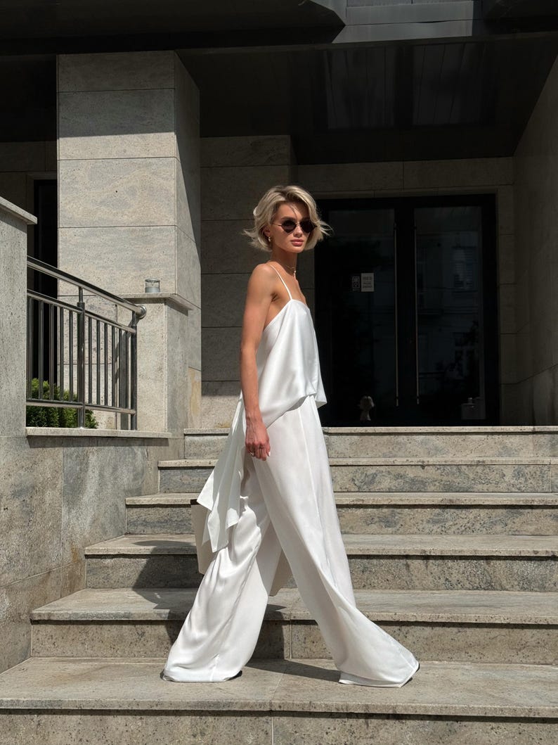 以下が含まれることがあります： 白いサテンのツーピースの衣装。キャミソールとワイドパンツで構成されています。短いブロンドの髪とサングラスをかけた人が、石段を歩いています。この衣装は、流れるようなエレガントな外観をしています。