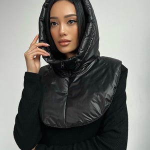 May include: Black hooded puffer scarf with a high collar and a zip closure. The scarf is made of a shiny, quilted material and is designed to be worn over the shoulders. The model is wearing a black sweater.