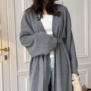 Könnte beinhalten: Eine langärmelige graue Strickjacke mit offener Vorderseite, einem passenden Stoffgürtel in der Taille und überschnittenen Schultern. Die Strickjacke wird über einem weißen gerippten Tanktop und einer hellblauen Jeans getragen, ergänzt durch eine beige geflochtene Handtasche.