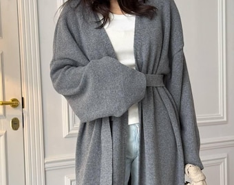May include: A long-sleeved gray knit cardigan featuring an open front, a matching fabric tie belt at the waist, and dropped shoulders. The cardigan is worn over a white ribbed tank top and light blue denim jeans, paired with a woven beige handbag.