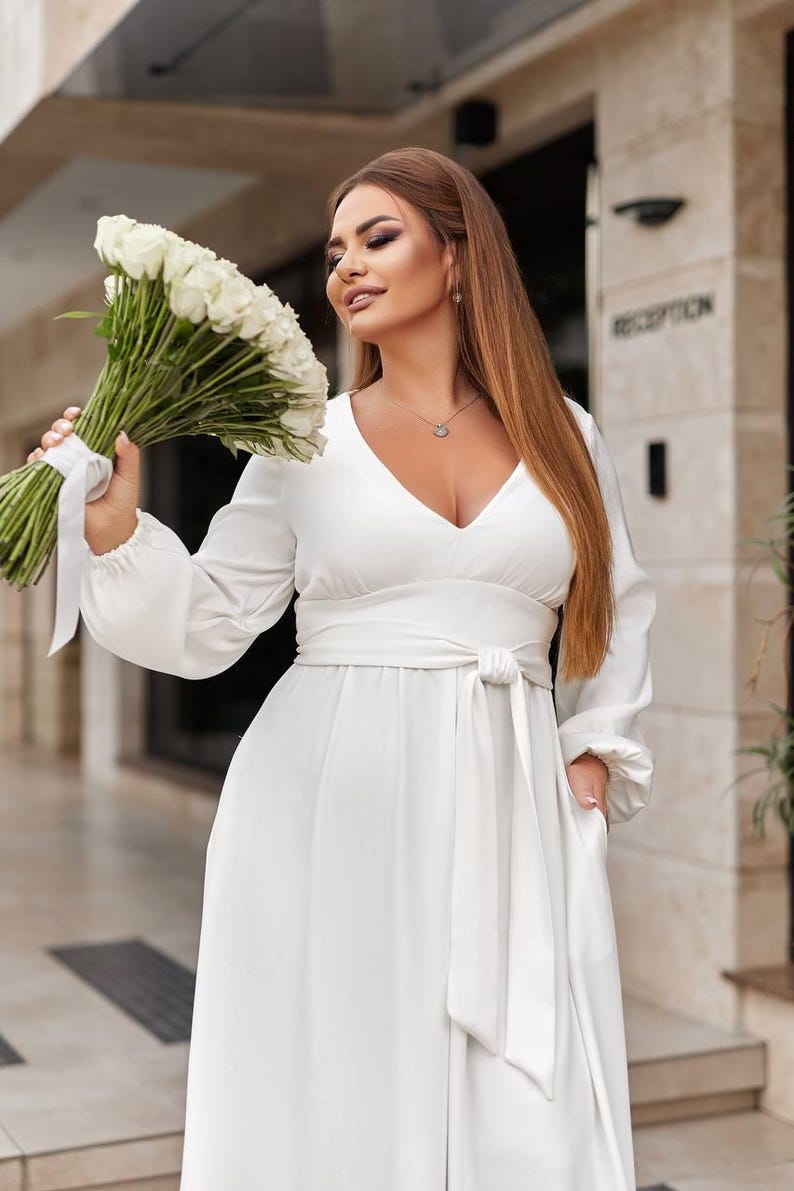 May include: A white, long-sleeved dress with a V-neck and a tie-waist. The dress features a flowing skirt and pockets. The person is holding a bouquet of white roses. The background is a building with the word "RECEPTION".