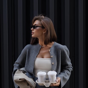 May include: A charcoal gray knit cardigan with a button-down front and matching knit skirt, paired with a white tube top, a beige leather handbag, and two white disposable coffee cups held in a cardboard carrier, worn with dark sunglasses.